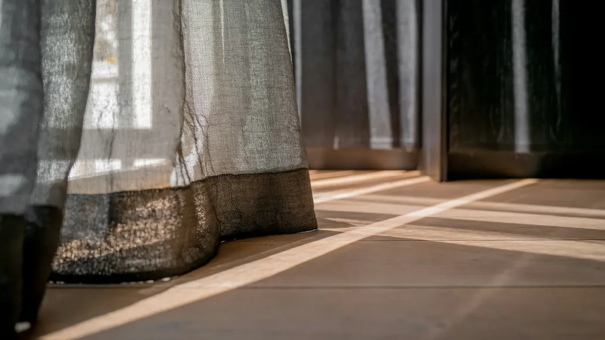 Una tenda chiara davanti a una portafinestra, con la luce del sole che disegna una striscia sul pavimento.